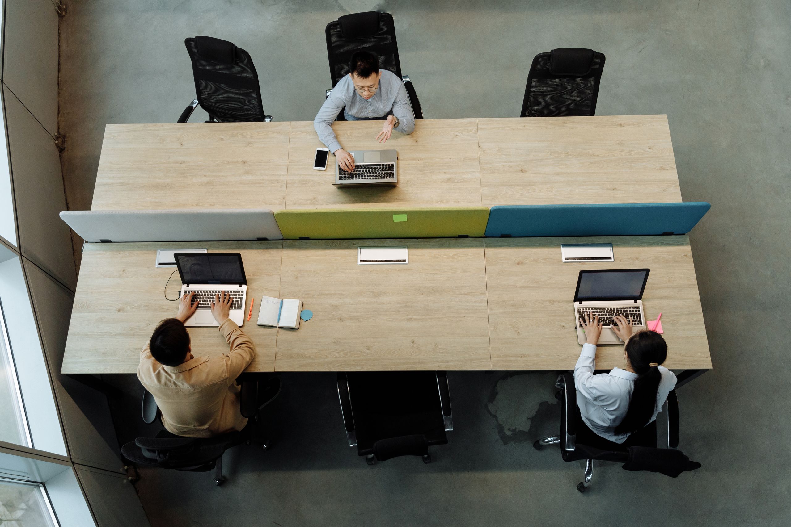 Three colleagues working at a shared office desk, using laptops in a modern workspace.