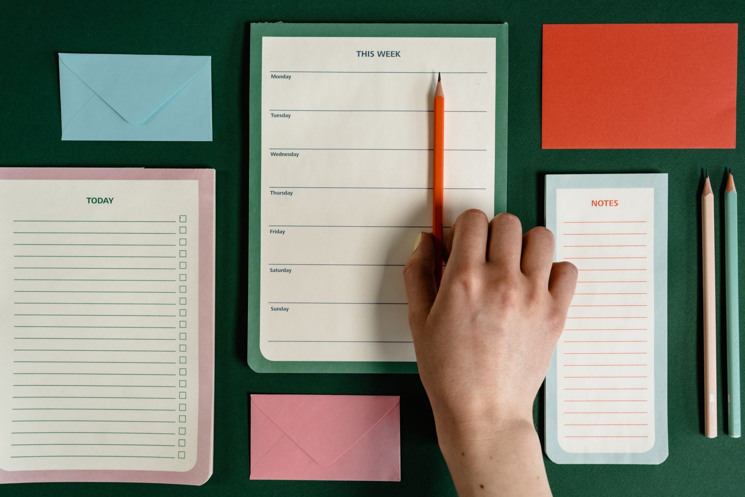Top view of a hand using a notepad surrounded by colorful stationery on a green surface.